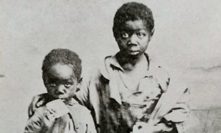 Old photo of two young black slave boys dressed in tattered clothing.