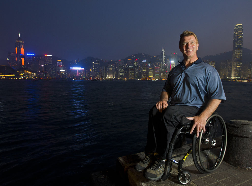 On voit ici un Rick Hansen souriant dans son fauteuil roulant, devant un cours d'eau et la silhouette d'une ville en arrière-plan.