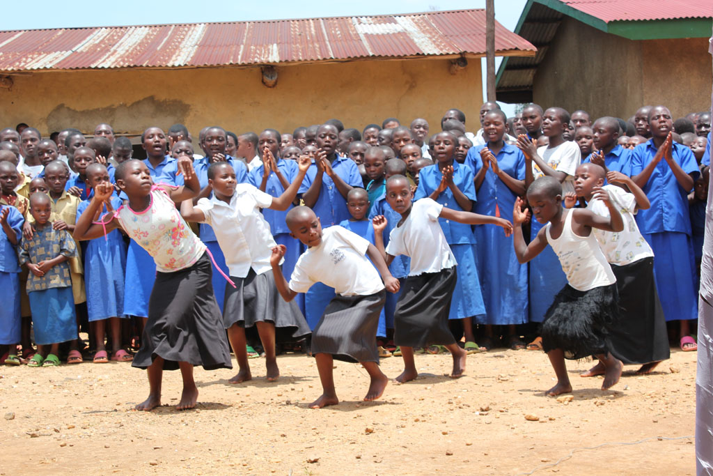 A group of Rwandan school children are dancing, while a larger group behind them looks on and claps.